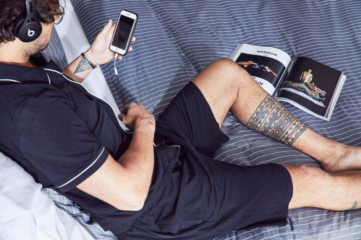 Person in pajamas listening to music on a smartphone while reading a magazine in bed.