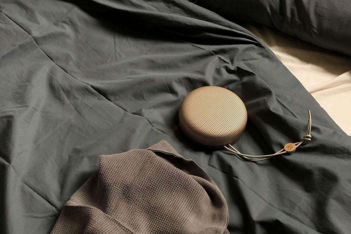 Round portable speaker on rumpled dark grey bedding beside a brown waffle knit throw