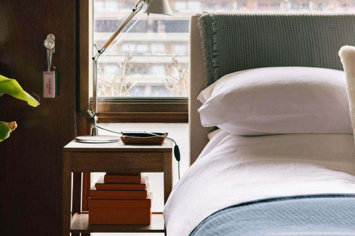 Close up of a bed with white bedding, a pale blue blanket and a green headboard by a window