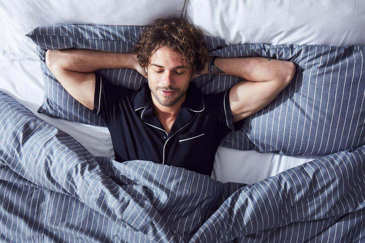 Man in navy pyjamas lying with his hands behind his head in navy and white striped bedding
