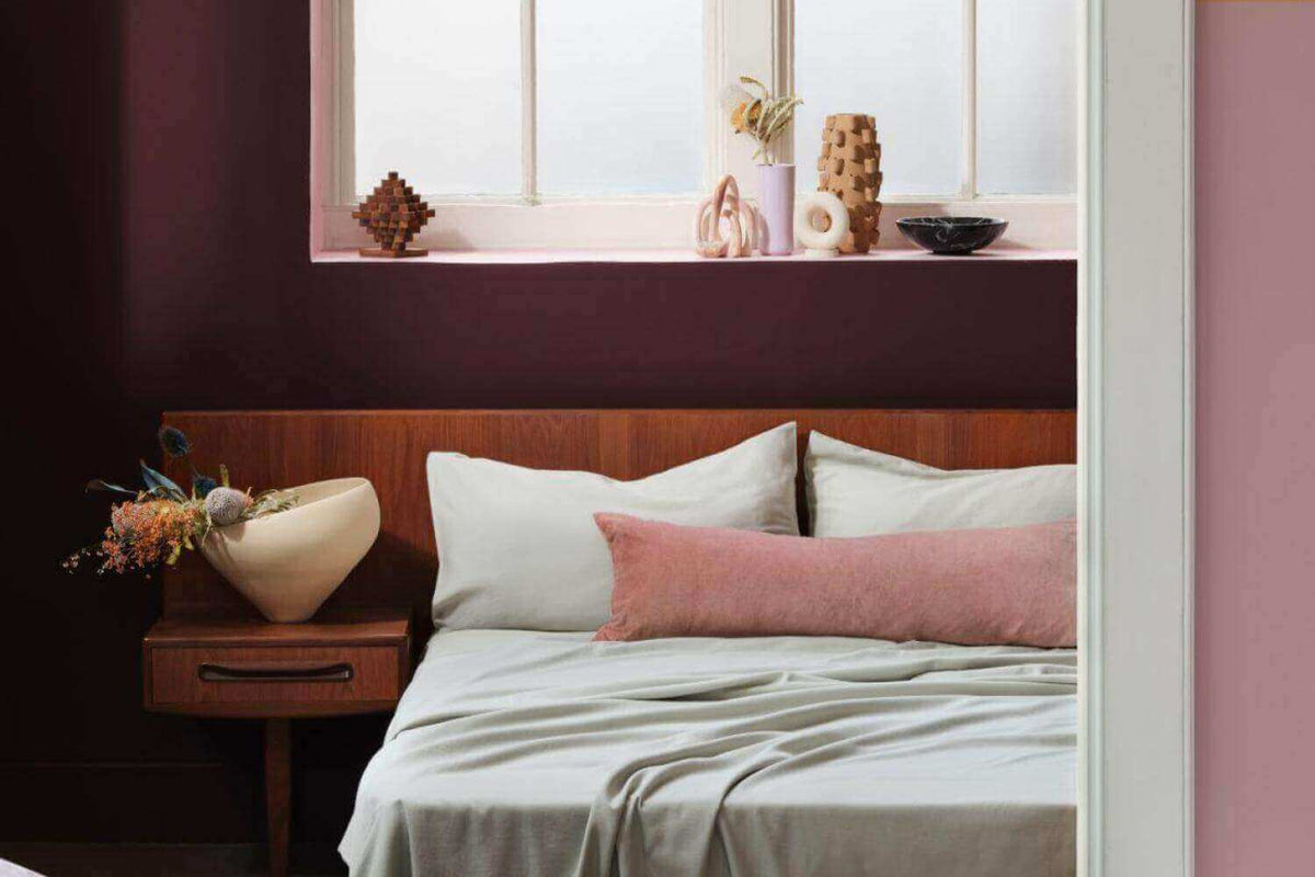 Minimal bedroom with light grey bedding, a pink cushion, a wooden headboard and a plum wall