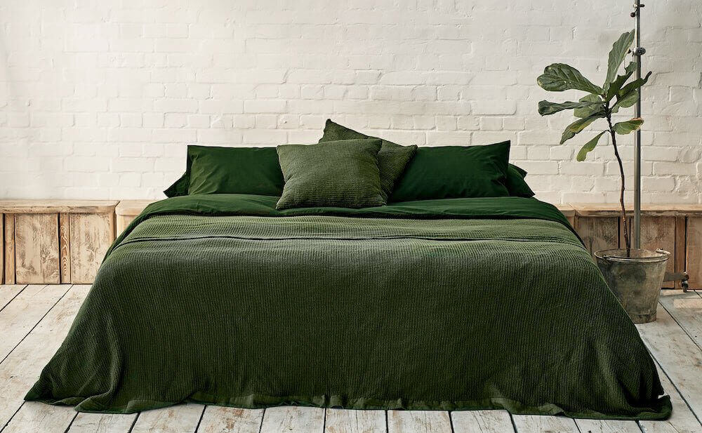 Bed dressed in dark green bedding with matching pillowcases against a white brick wall