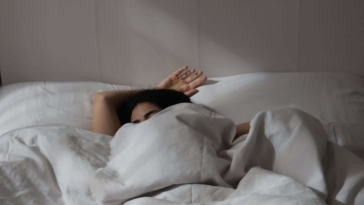 Woman stretching her arm out from under rumpled white bedding in a softly lit bedroom