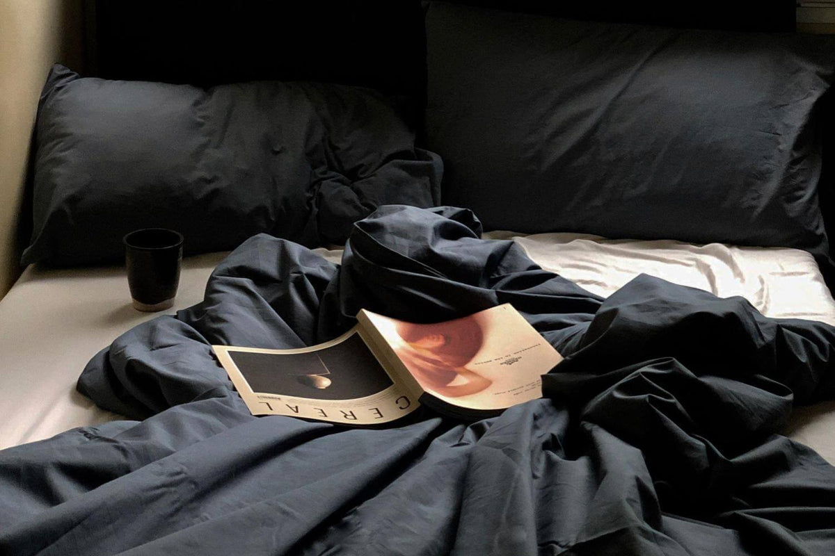Rumpled navy bedding on an unmade bed with an open magazine and a dark glass