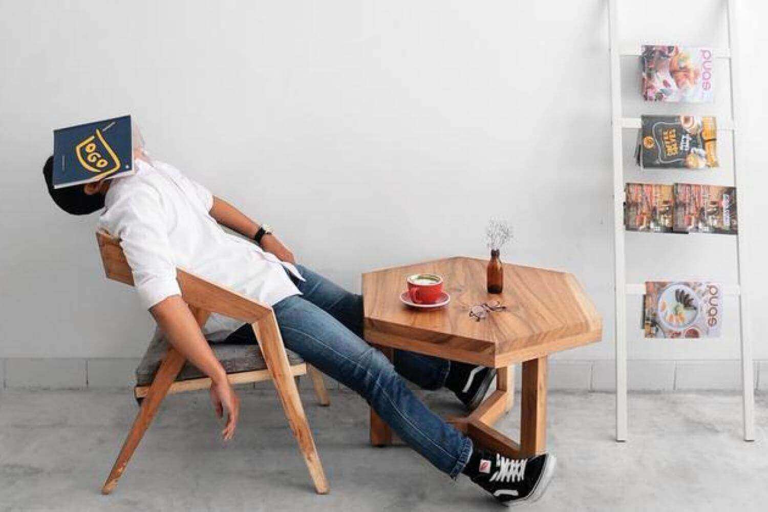 Person relaxing at a modern wooden table with a coffee cup, humorously resting with a book over their face.