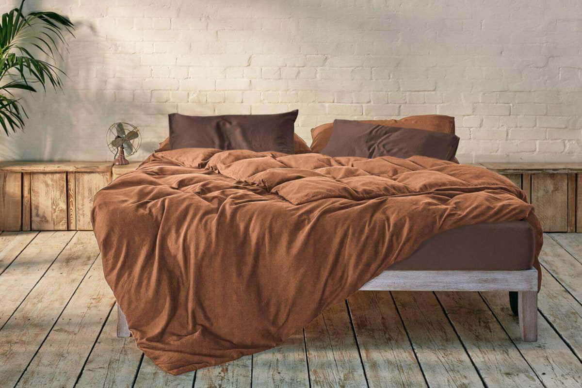 Low bed with rumpled terracotta bedding on a weathered wooden floor against a white brick wall