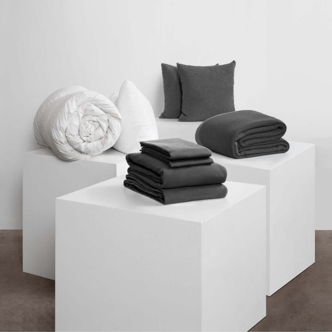 Neatly arranged bedding items including blankets, pillows, and cushions on white pedestals for spring cleaning inspiration.