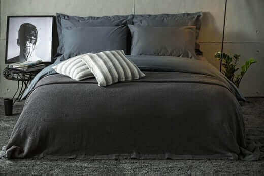 Bed with charcoal bedding, a striped cushion and a knitted throw against a grey wall